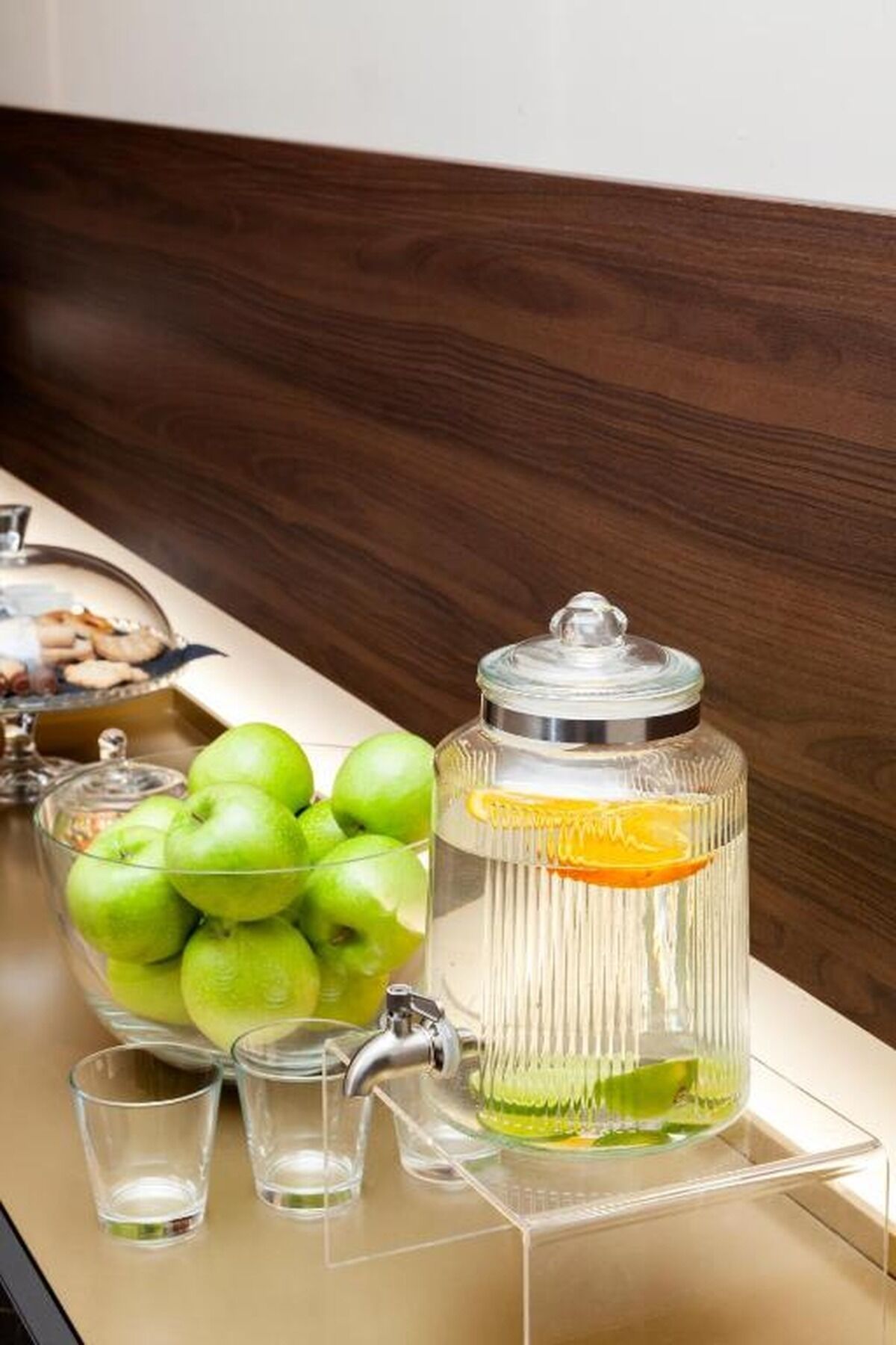 A kitchen counter with a glass jar containing a beverage, a bowl of green apples, and two shot glasses.