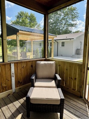 A covered outdoor seating area with a cushioned chair and a small table, wooden floor, and a view of a building and trees through large windows.
