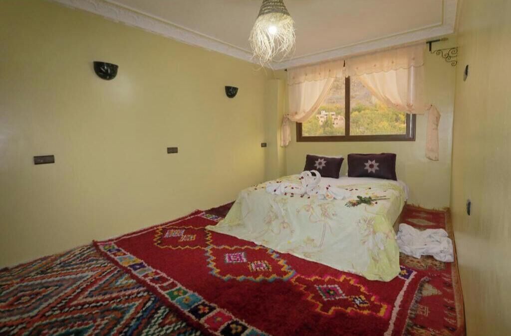 A bedroom with a bed, a window with curtains, a hanging light fixture, and a patterned rug.