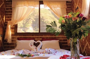 Una cama con una toalla dispuesta en forma de corazón, un florero con flores y una vista de un paisaje montañoso a través de una ventana.