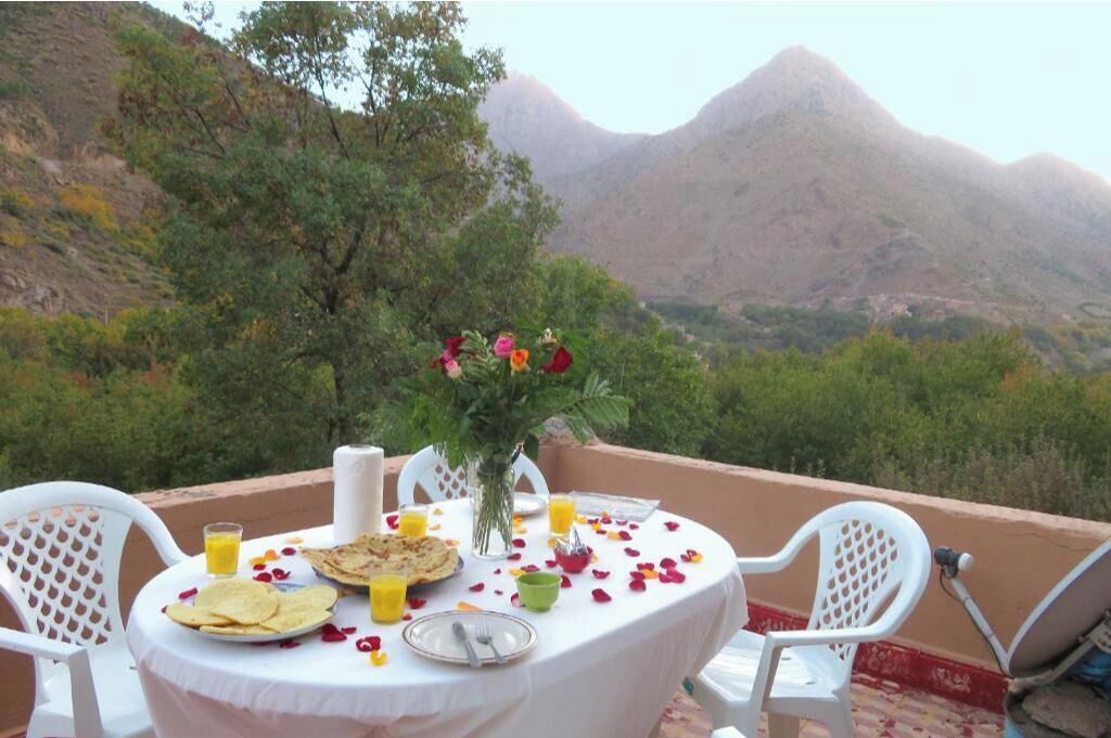 A table set for breakfast with a mountain view, a vase of flowers, and scattered rose petals.