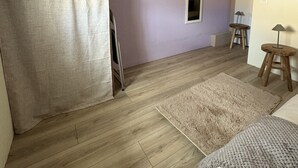 A room with a purple wall, wooden flooring, a bed, a mirror, a wardrobe, and a small stool.