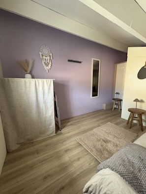 Una habitación con pared morada, suelo de madera, cama, espejo, armario y un taburete pequeño.