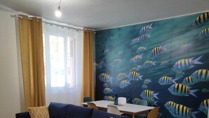 Un espace de vie comprenant un canapé, une petite table ronde, une table à manger avec des chaises et une fresque murale représentant des poissons.