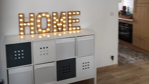 Un meuble de rangement blanc sur lequel est inscrit le mot « MAISON » en lettres lumineuses, un appareil de climatisation placé au-dessus, et un coin cuisine en arrière-plan.