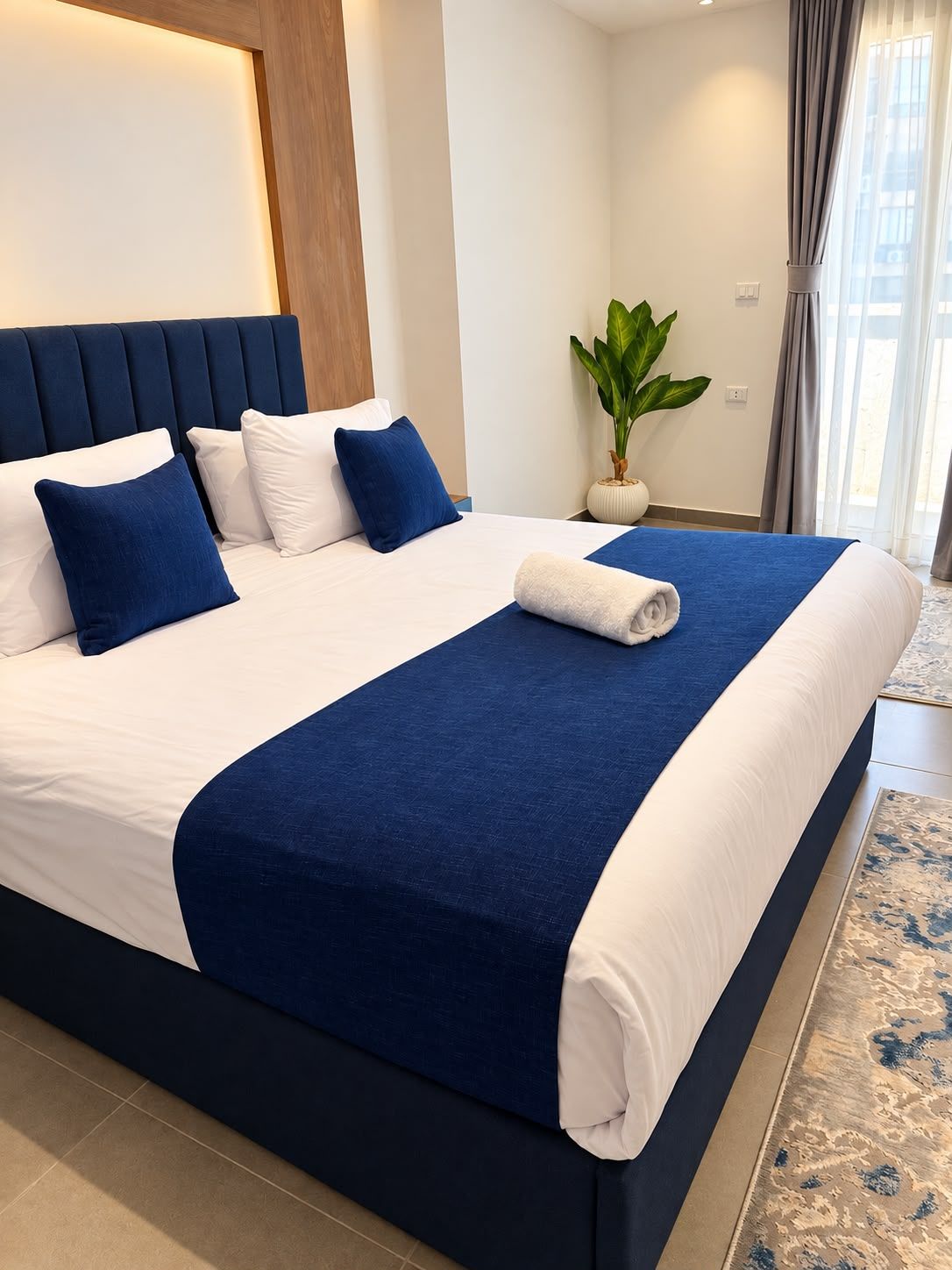 Ein ordentlich bezogenes Bett mit blauer und weißer Farbgebung, ein Blumentopf und ein Blick ins Freie durch ein Fenster.
