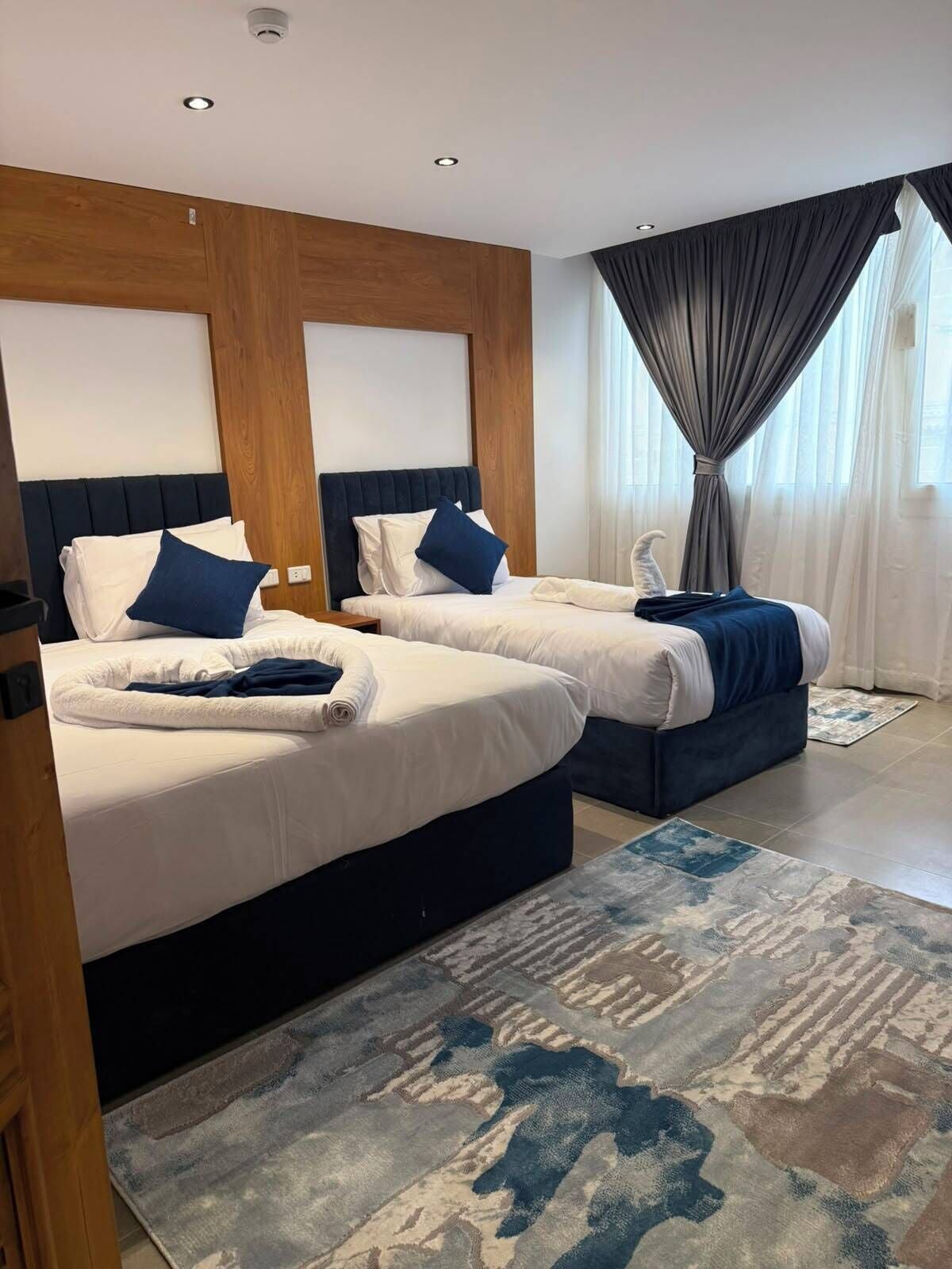 A hotel room with two beds, a large window with curtains, and a patterned rug.