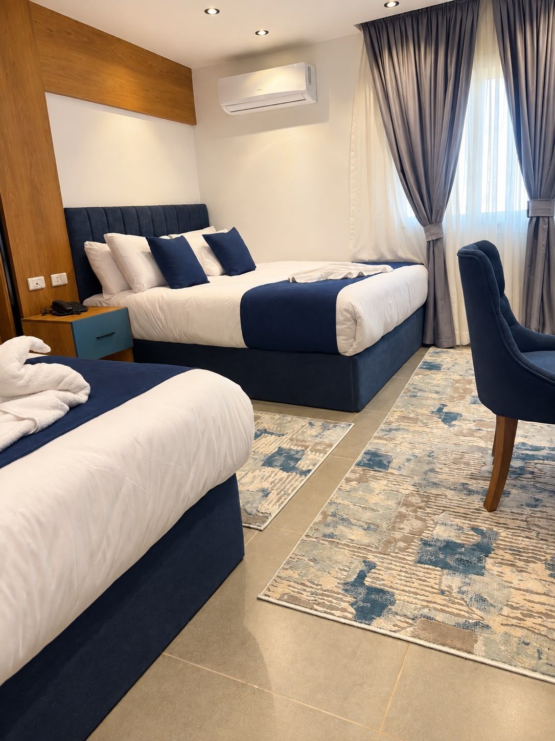 Habitación de hotel con dos camas, un escritorio, una silla, un equipo de aire acondicionado y una ventana con cortinas.