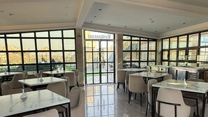 Un amplio comedor con varias mesas y sillas, ventanas grandes que ofrecen vistas de la ciudad y un diseño de techo moderno.
