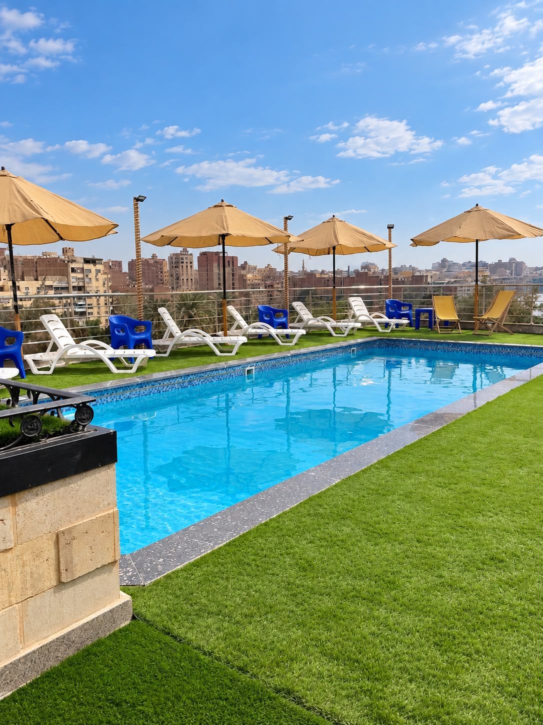A swimming pool with lounge chairs and umbrellas, surrounded by a grassy area and a cityscape in the background.