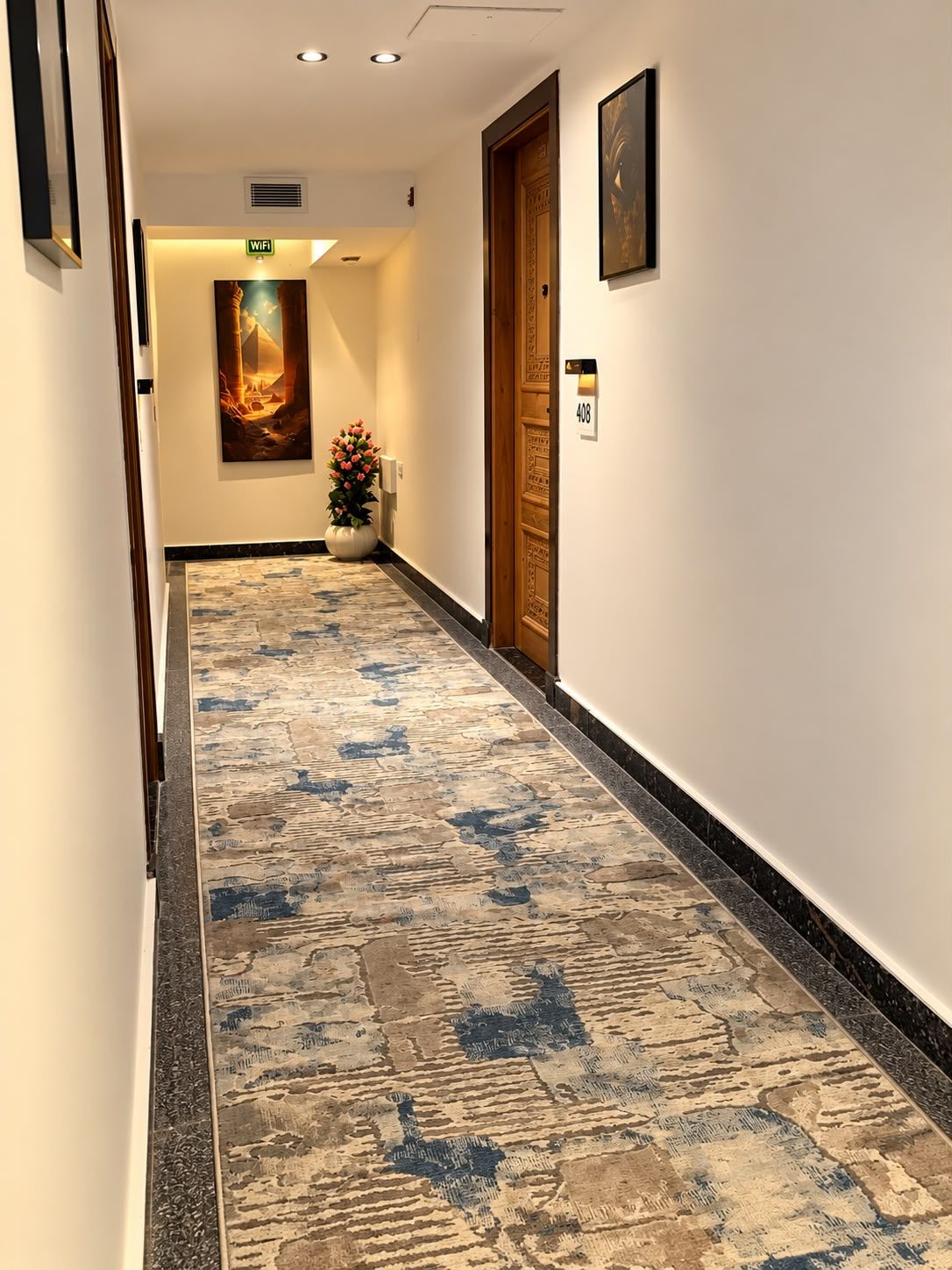 Un couloir d’hôtel avec un tapis à motifs, une porte en bois, un tableau encadré et un panneau indiquant la disponibilité du Wi-Fi.