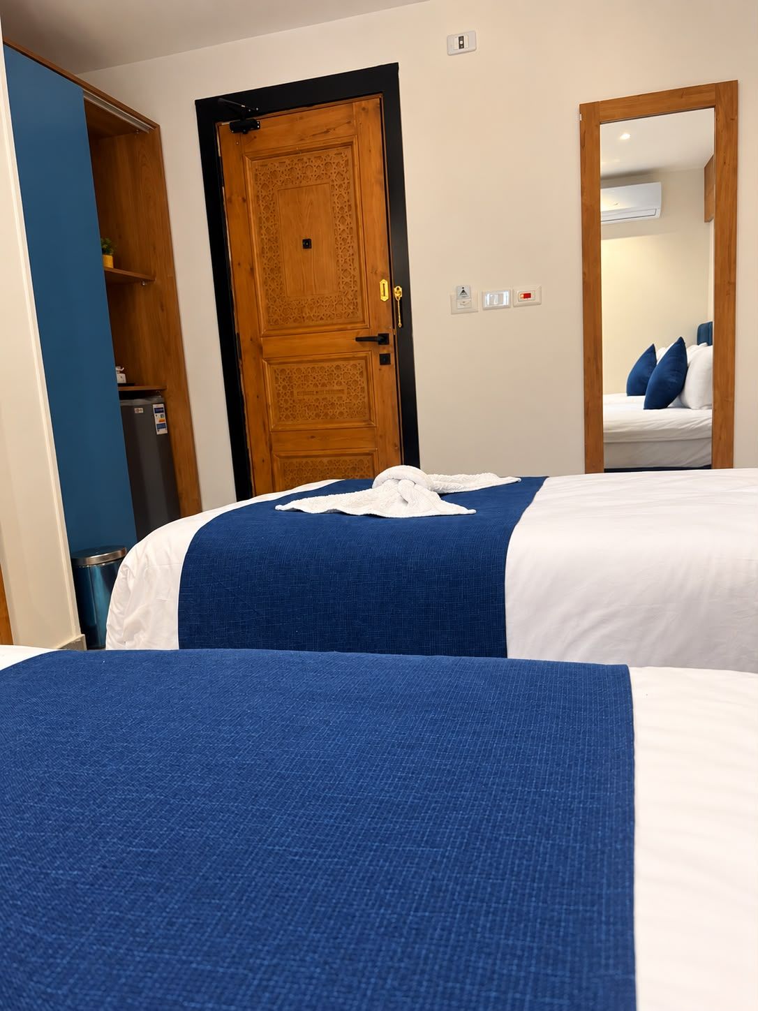 Quarto de hotel com duas camas, porta de madeira, espelho e pequena cozinha.