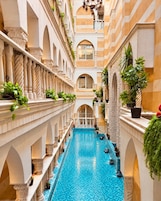 A luxurious indoor swimming pool with multiple levels of balconies, surrounded by classical architecture and potted plants.