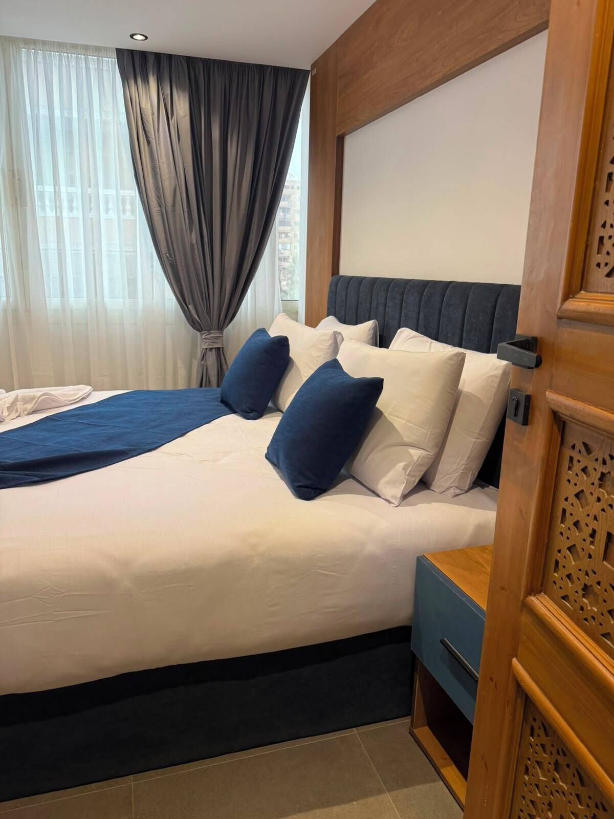 A hotel room with a bed, a nightstand, a wooden panel, and a window with curtains.