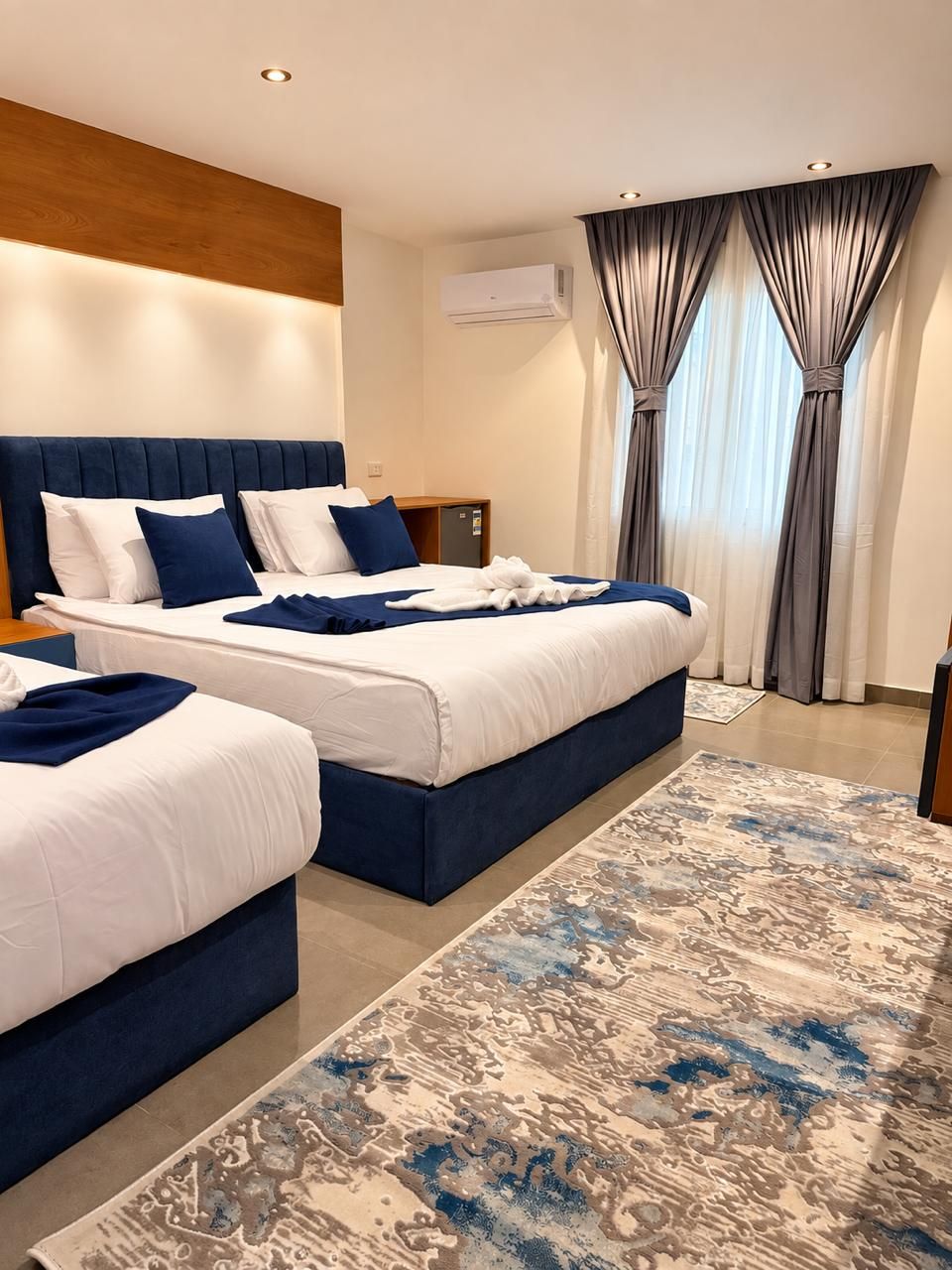 A hotel room with two beds, a large window with curtains, a wooden headboard, and a modern air conditioning unit.