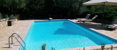 Une piscine rectangulaire bordée de pierres, entourée de chaises longues et d’un grand parasol.