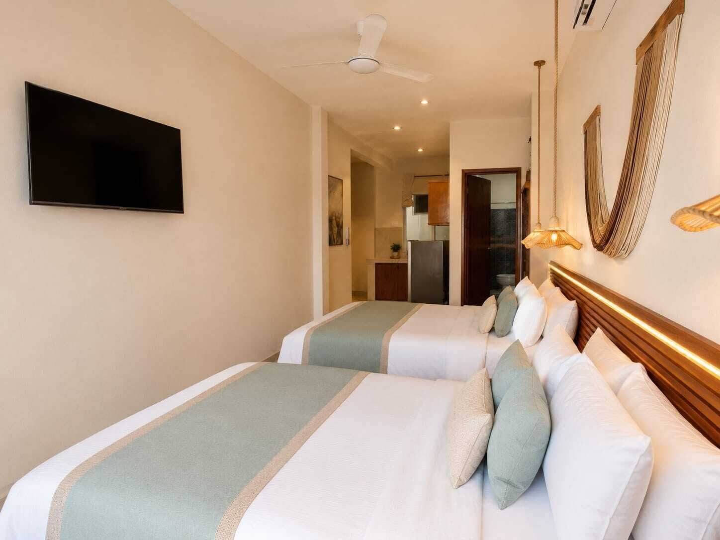 A hotel room with two beds, a flat-screen TV, a ceiling fan, and a kitchen area visible through an open door.