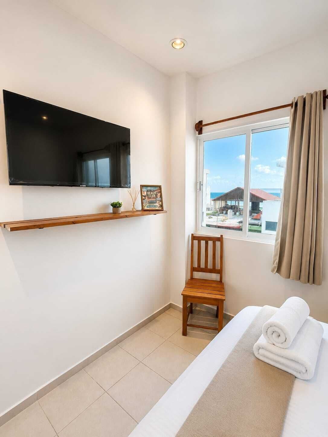 A hotel room with a bed, a TV mounted on the wall, a window with curtains, a chair, and a small shelf with a picture and a plant.
