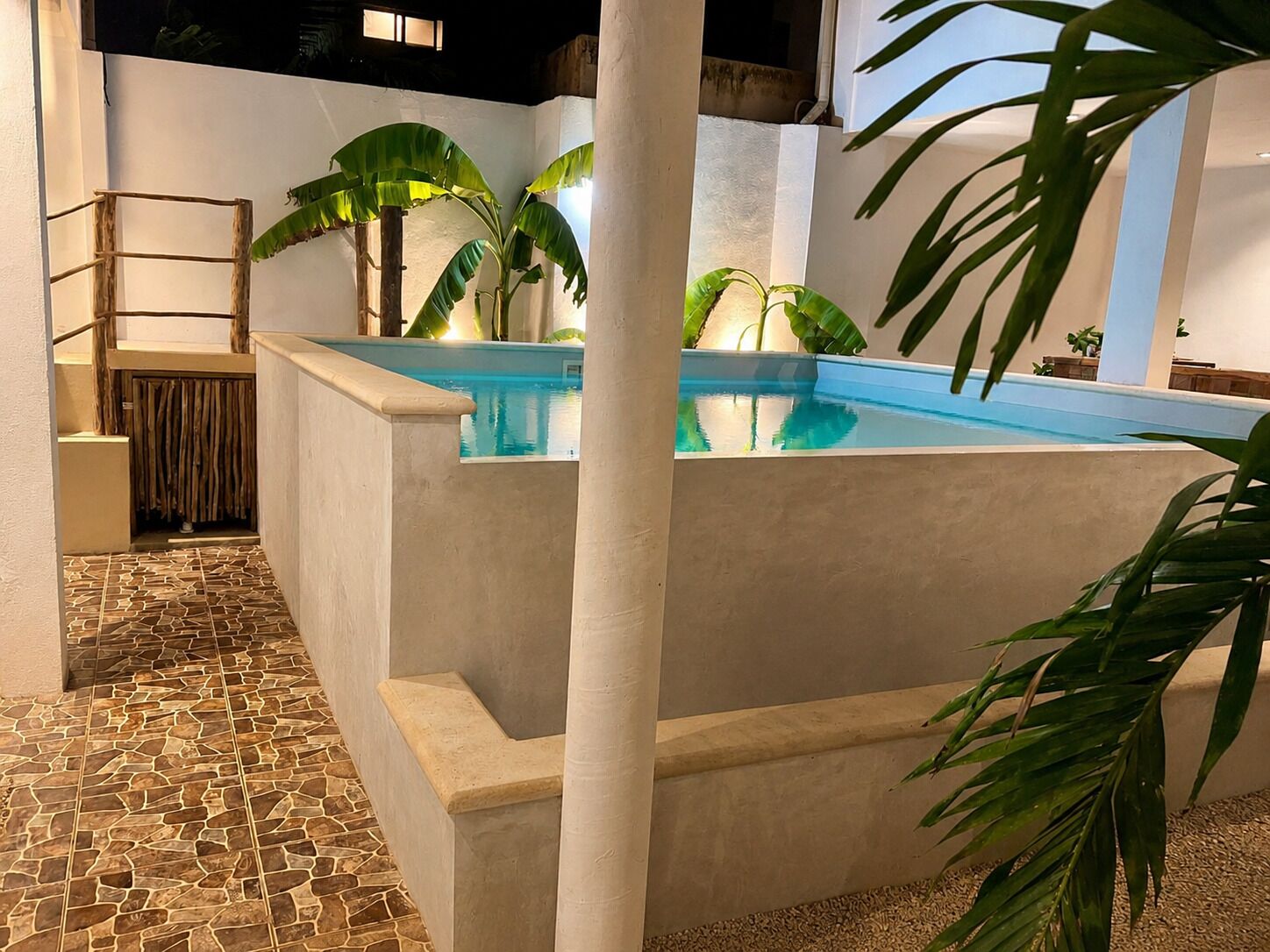Uma piscina com piso de ladrilho de pedra, rodeada por paredes brancas e grandes plantas verdes.