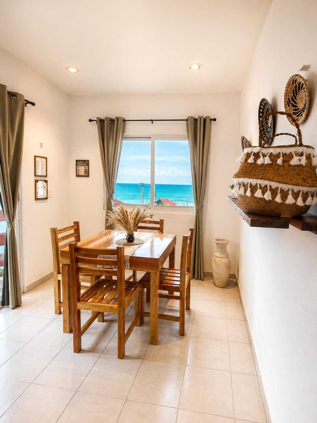 A dining area with a wooden table and chairs, a view of the sea through a window, and a woven basket on a shelf.