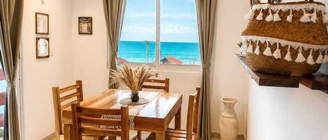 A dining area with a wooden table and chairs, a view of the sea through a window, and a woven basket on a shelf.