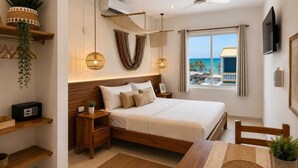 A modern bedroom with a bed, bedside tables, a TV, a ceiling fan, and a window with a view of the beach. - Hotel Isla Mujeres Centro (Isla Mujeres)