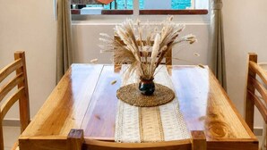 A wooden dining table with a woven placemat, two chairs, and a vase with dried pampas grass. A window with beige curtains offers a view of the ocean and a building with a red roof. - Hotel Isla Mujeres Centro (Isla Mujeres)