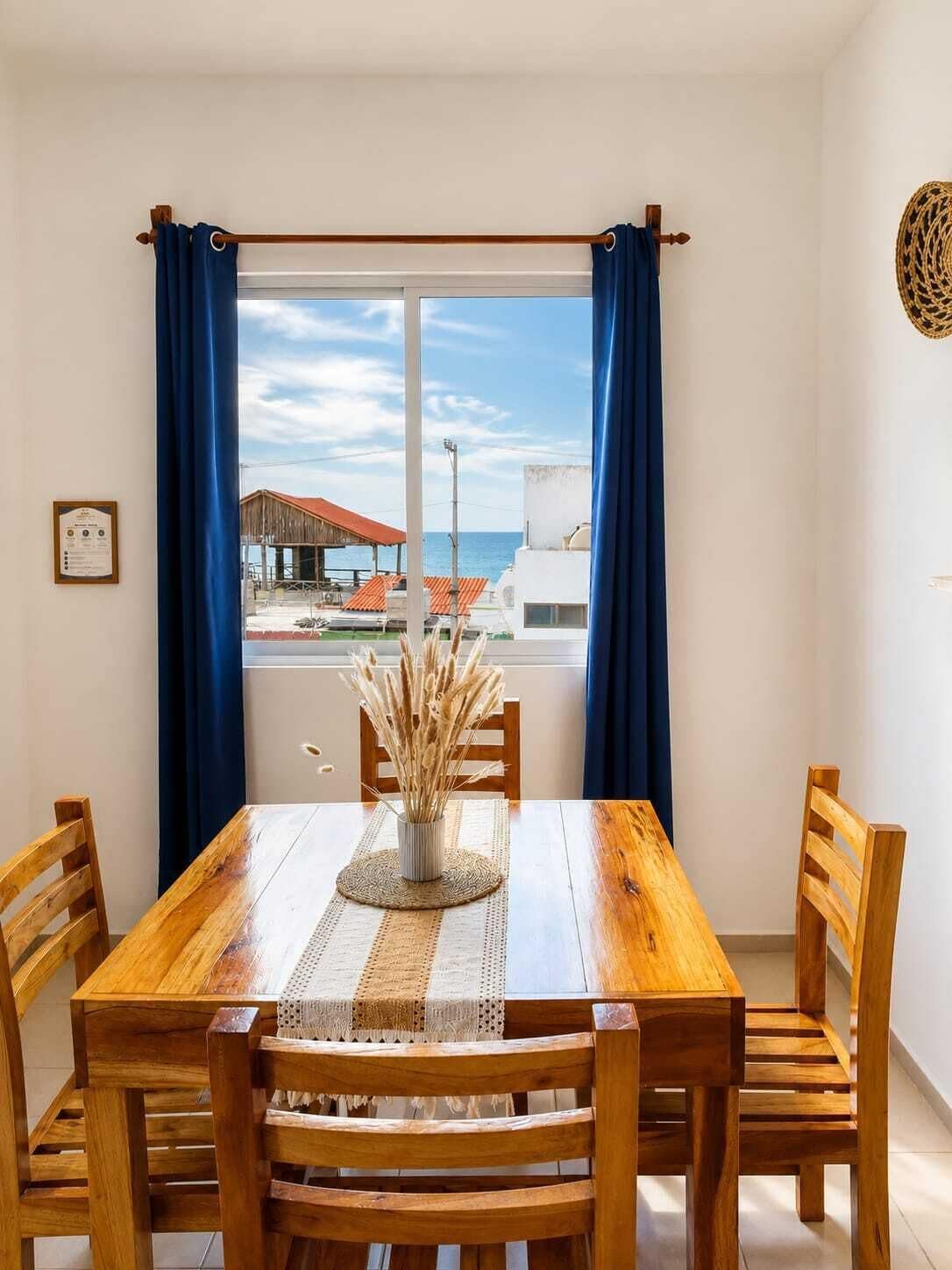 A dining area with a wooden table and chairs, a view of the sea, and a building with a red roof.