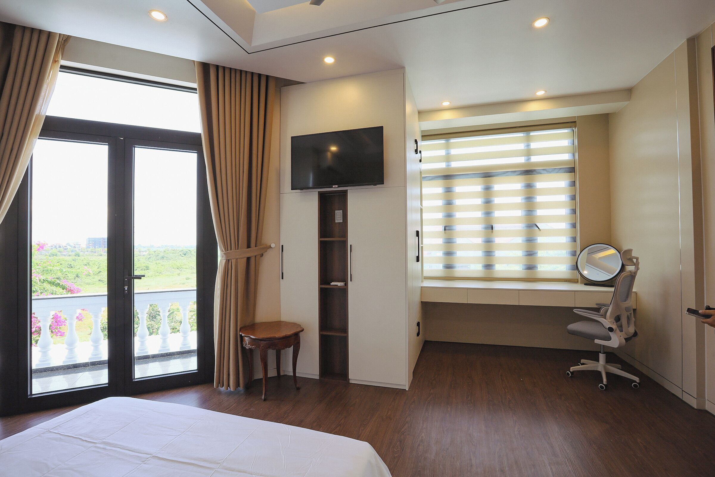 Een hotelkamer met een bed, een televisie, een bureau met een stoel, een raam met jaloezieën en een balkon met uitzicht.