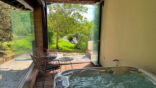 Ett bubbelbad med en träaltan och utemöbler, en glasdörr som leder ut till en trädgård och en fri utsikt över träd och en väg.