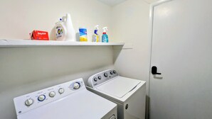 A laundry room with two washers and dryers, a shelf with cleaning supplies, and a door.
