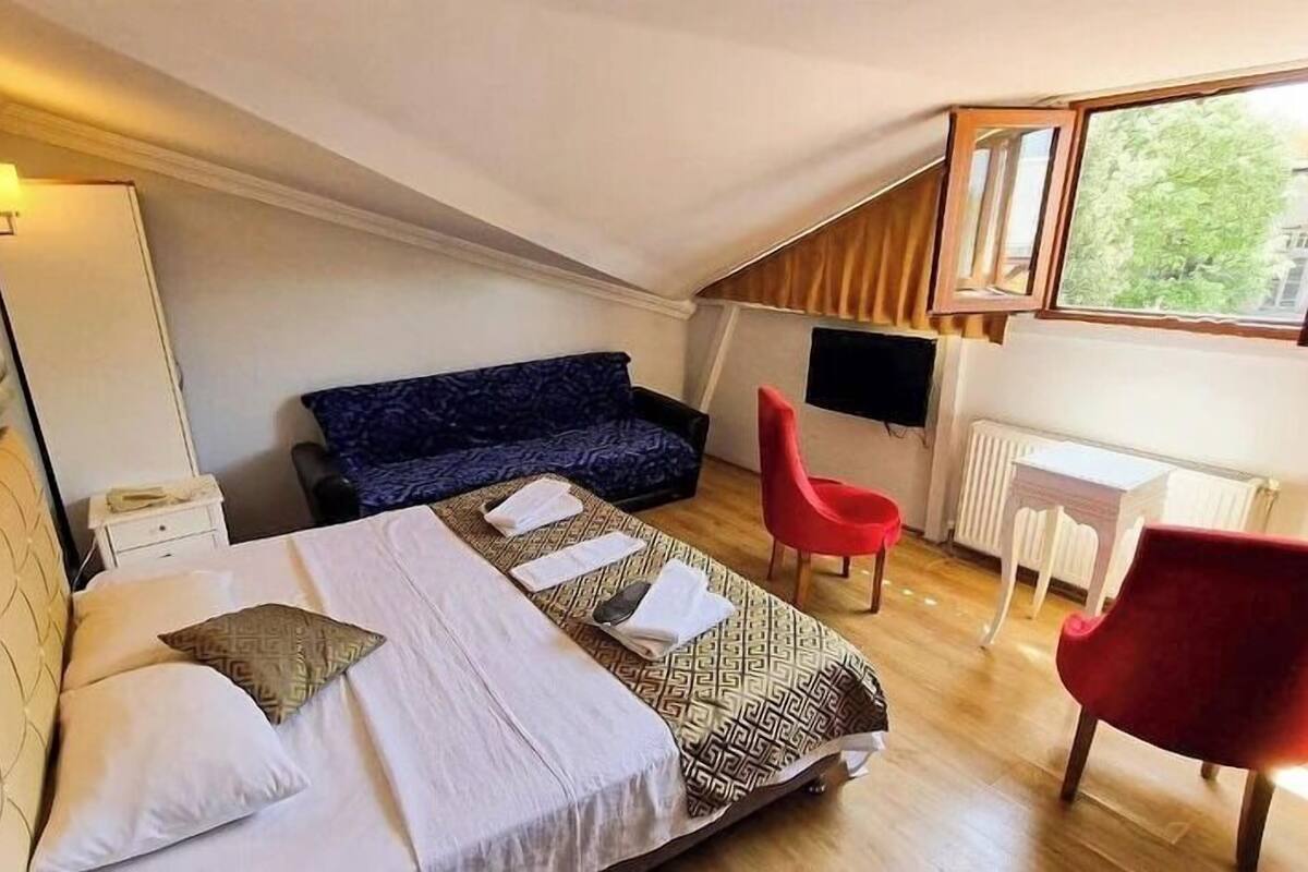 Un dormitorio con cama, una silla roja, sofá, escritorio y ventana.