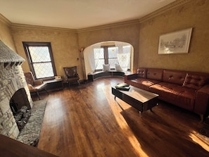 A living room with a stone fireplace, wooden flooring, a leather sofa, and a large window with a view. - Historic Holton House Spacious 2 bedroom Victorian with Charm and Home Bar (Milwaukee)