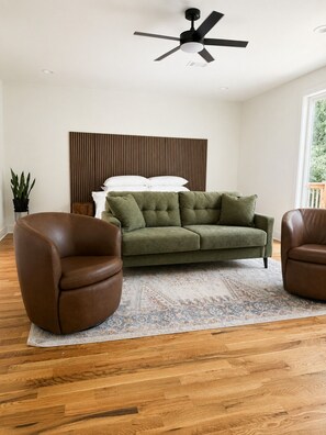 A modern living room with a green sofa, two brown armchairs, a ceiling fan, and a wooden floor.