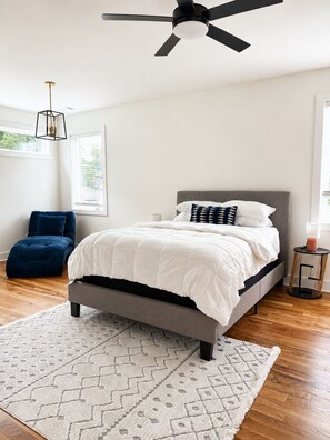 A bedroom with a bed, a ceiling fan, a window, a blue chair, and a patterned rug.