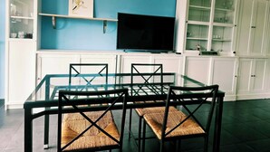 Un comedor con una mesa y sillas de vidrio, un televisor de pantalla plana, armarios blancos y un equipo de aire acondicionado.