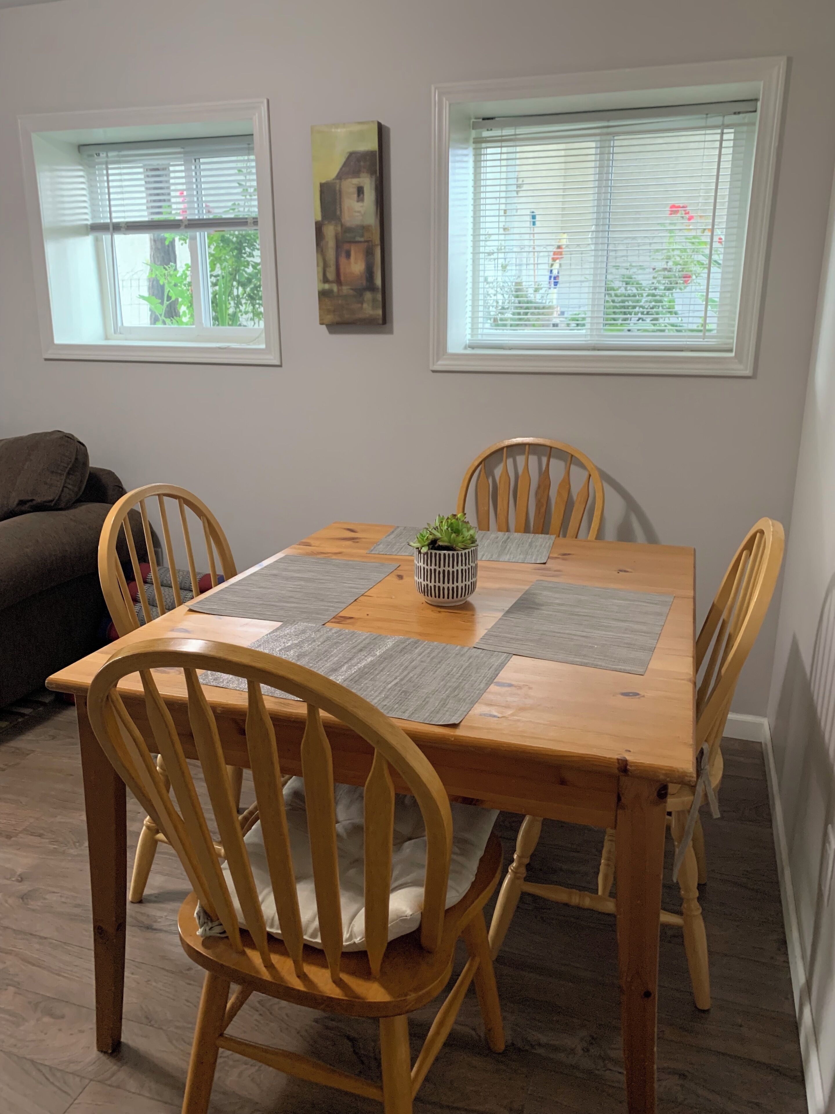 Un coin repas avec une table en bois, quatre chaises, deux fenêtres équipées de stores et un tableau accroché au mur.