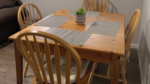 Een eetruimte met een houten tafel, vier stoelen, twee ramen met luiken, en een schilderij aan de muur.