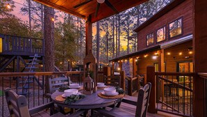 An outdoor dining area with a wooden deck, a stone fireplace, and a view of the surrounding forest. - 21-Ft Swim Spa Pool | 2 King Suites | Game Loft | Playset | Swings | Hot tub (Broken Bow)