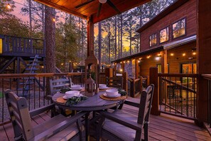 Un área de comedor al aire libre con terraza de madera, chimenea de piedra y vistas al bosque circundante.