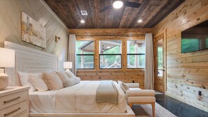 A wooden cabin bedroom with a bed, bedside tables, and a television. - 21-Ft Swim Spa Pool | 2 King Suites | Game Loft | Playset | Swings | Hot tub (Broken Bow)