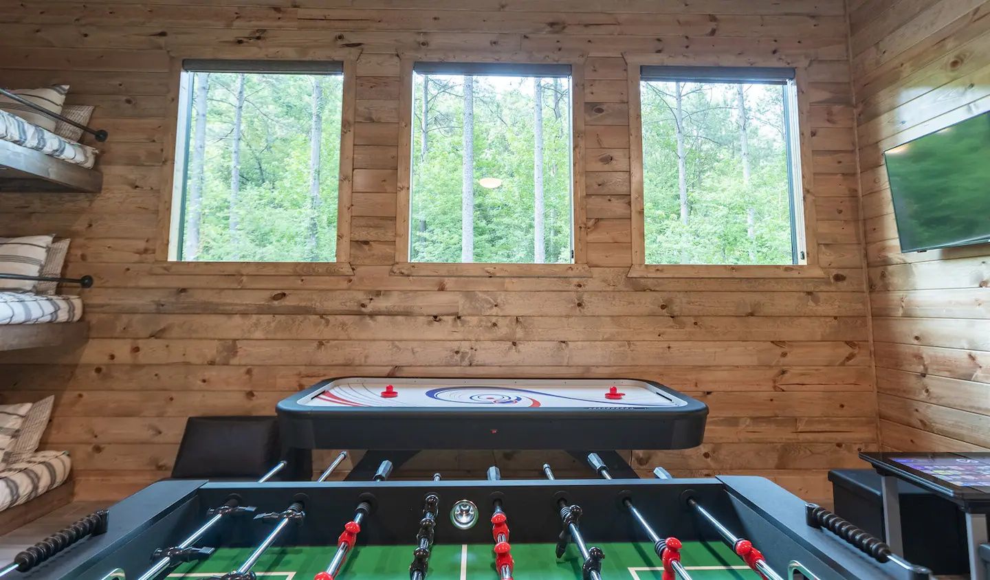 Una habitación con una mesa de fútbol de mesa, tres ventanas y paredes de madera.