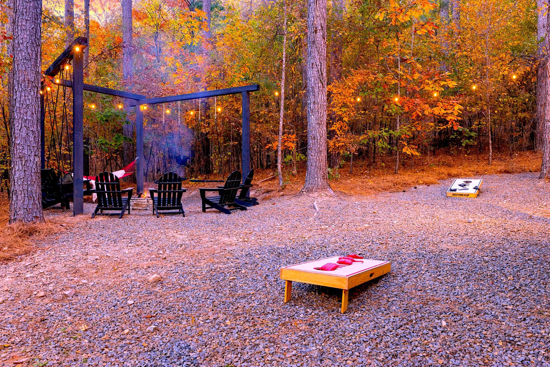 Un espacio de estar al aire libre rústico con sillas Adirondack, una hamaca y una mesa de madera con cojín rojo.