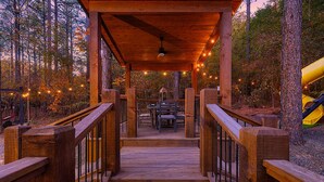 A wooden deck leading to a covered area with tables and chairs, surrounded by trees and a yellow slide. - 21-Ft Swim Spa Pool | 2 King Suites | Game Loft | Playset | Swings | Hot tub (Broken Bow)