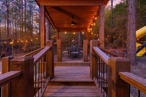 Una terraza de madera que conduce a un área cubierta con mesas y sillas, rodeada de árboles y un tobogán amarillo.