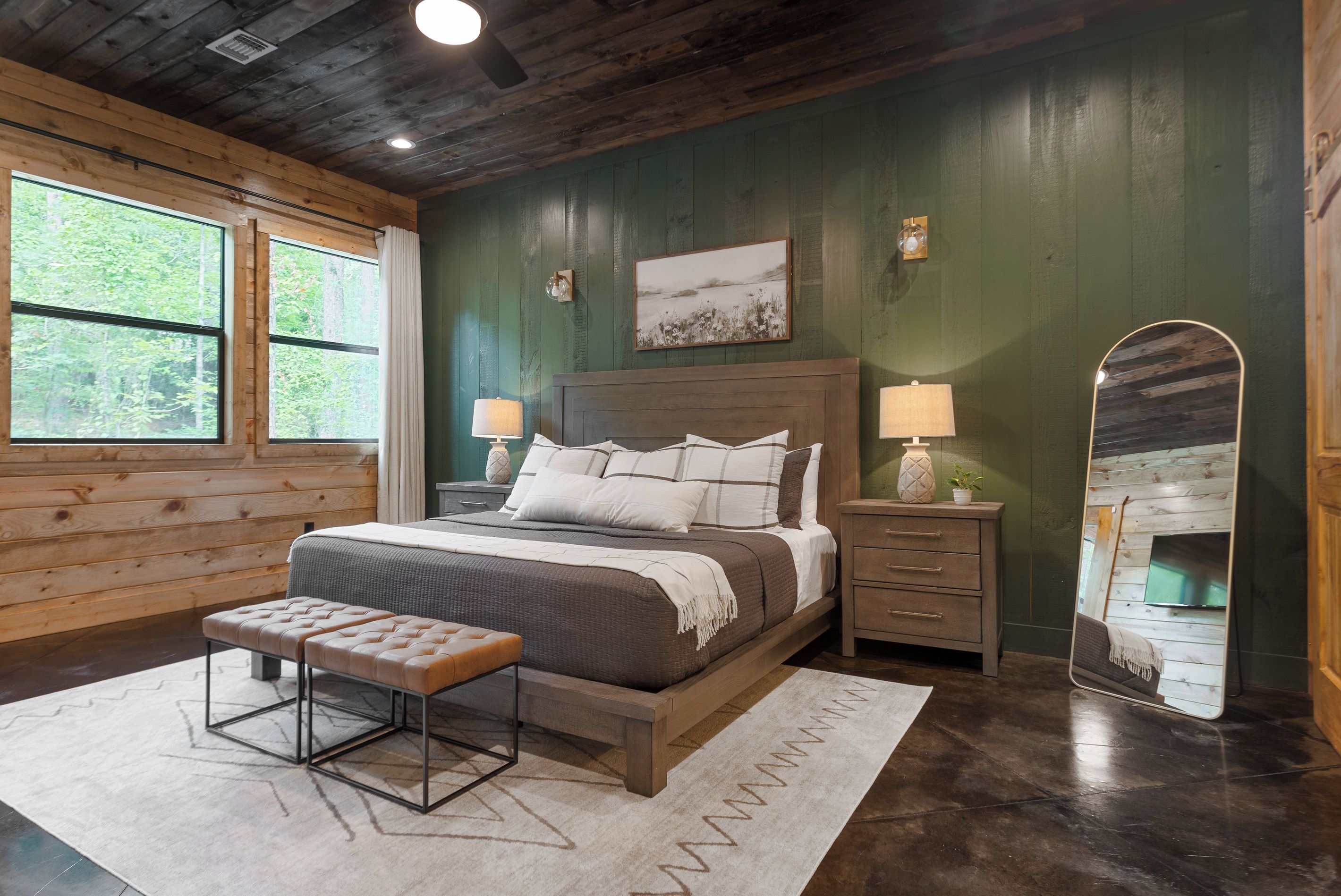 Une chambre à coucher avec un grand lit, des tables de chevet, un banc, un miroir et une fenêtre donnant sur des arbres.