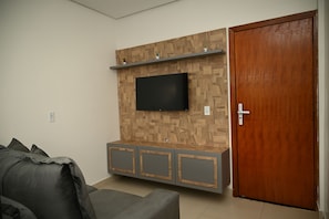 A room with a wooden wall panel, a flat-screen TV mounted above a grey cabinet, a wooden door, and a grey sofa.