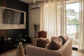 Una sala de estar con una televisión de pantalla plana, un sofá, una mesa de centro, una planta en maceta y vistas de edificios.
