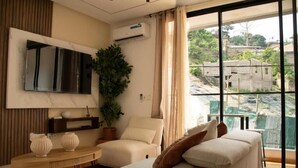A modern bedroom with a large window, a flat-screen TV, a sofa, a small table, and a potted plant. - LE DANORE VIP APPART hotel (Yaoundé)