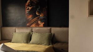 A bedroom with a bed, pillows, a bedside table, a wall-mounted painting, and a small shelf with plants. - LE DANORE VIP APPART hotel (Yaoundé)